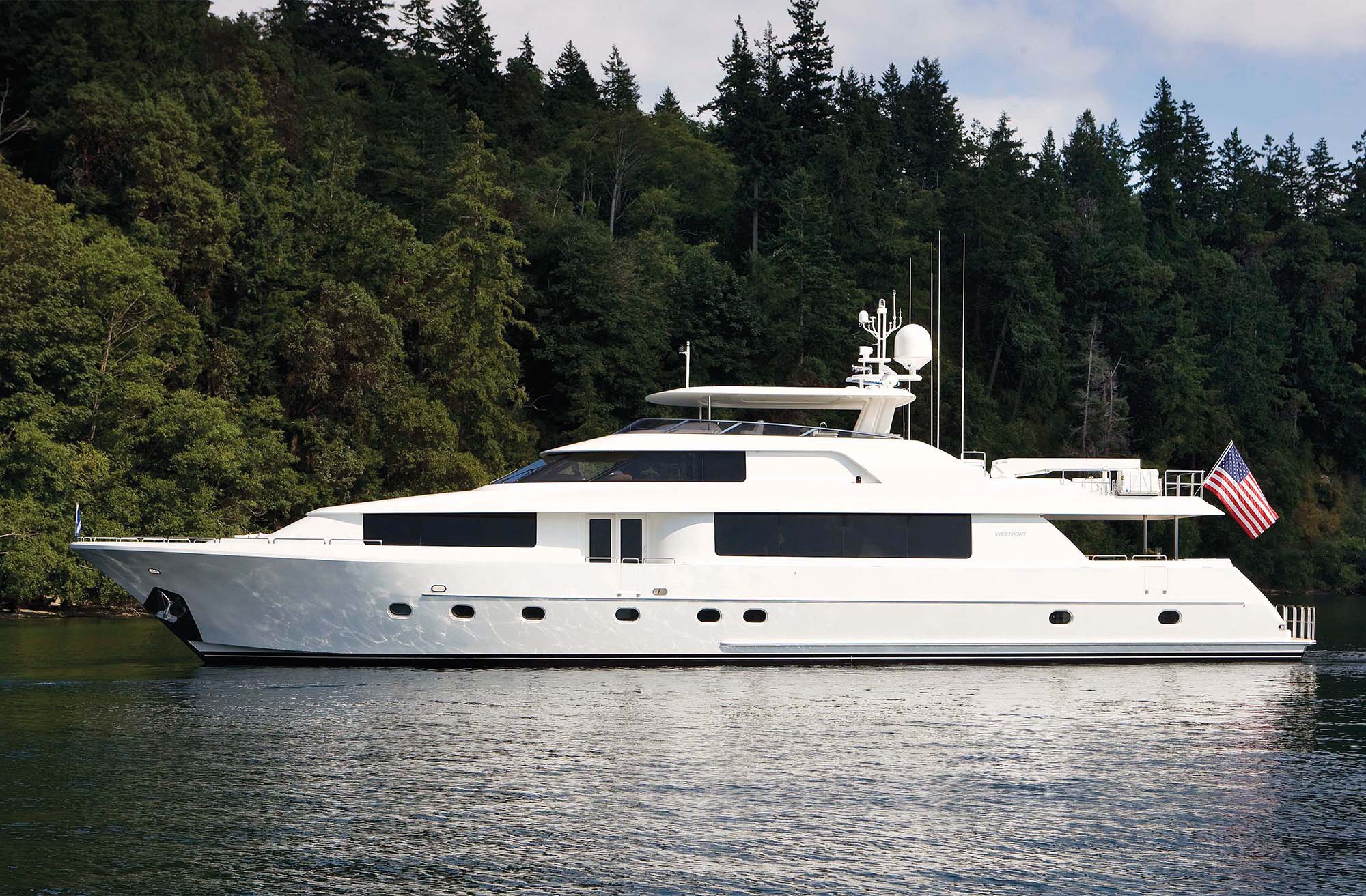 A white 112-foot yacht seen in profile with pine trees in the background