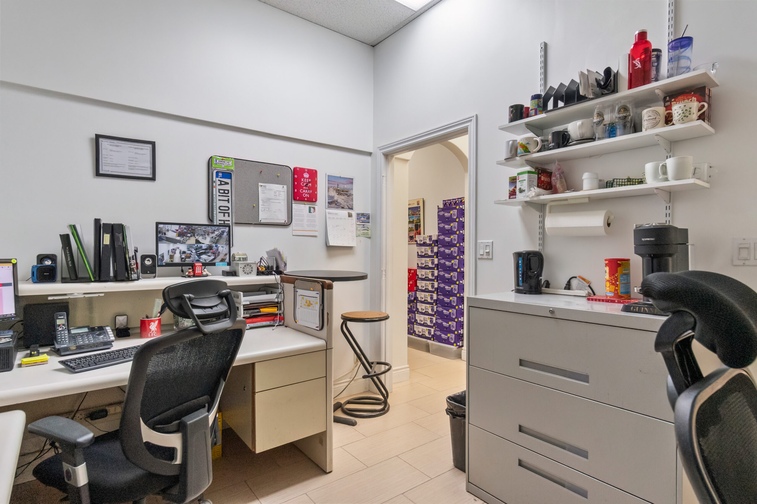 Office workspace with desks, shelves, and a doorway leading to another room