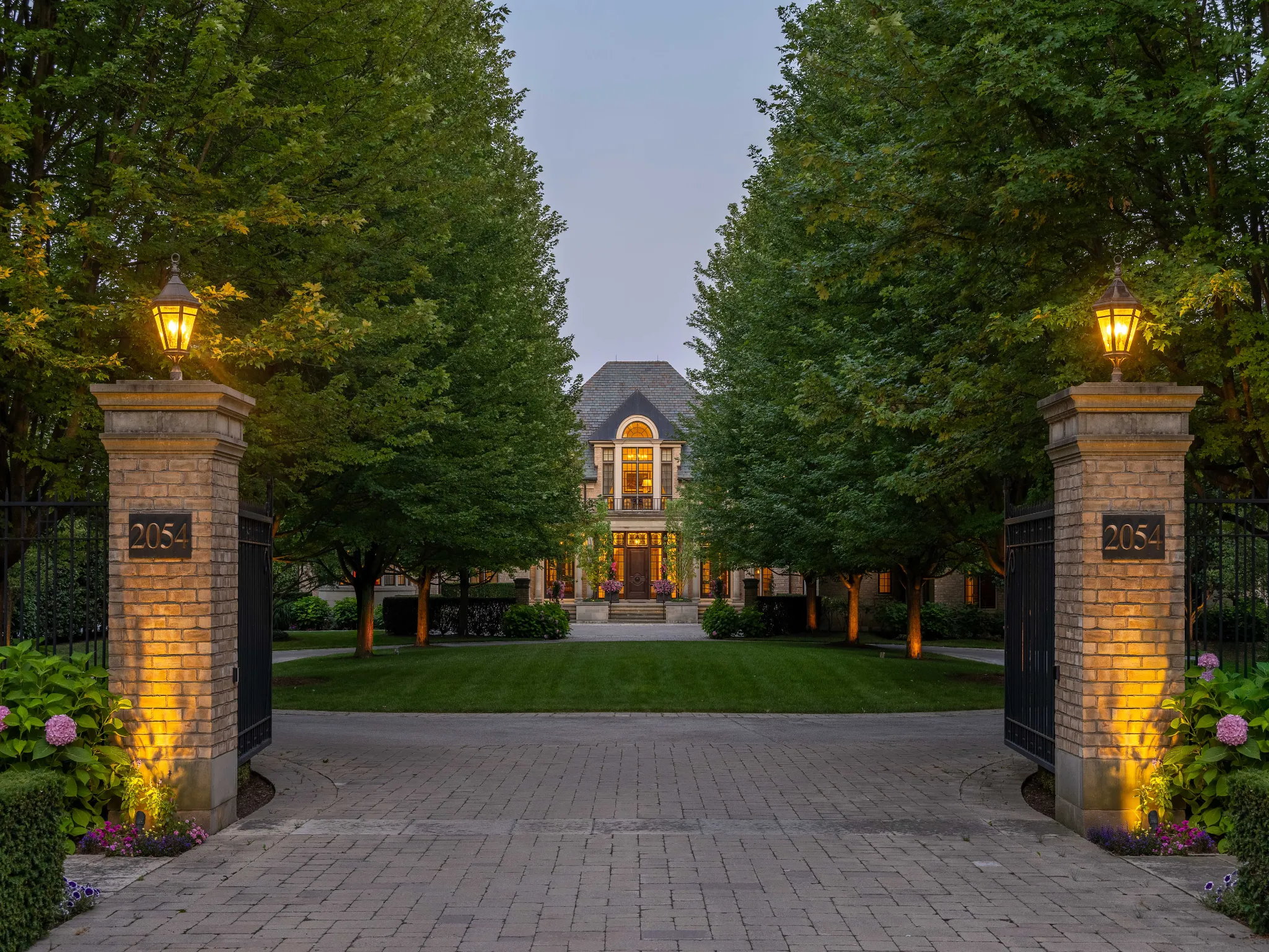 exterior of home flanked by large green mature trees and two gate pillars with lights on them