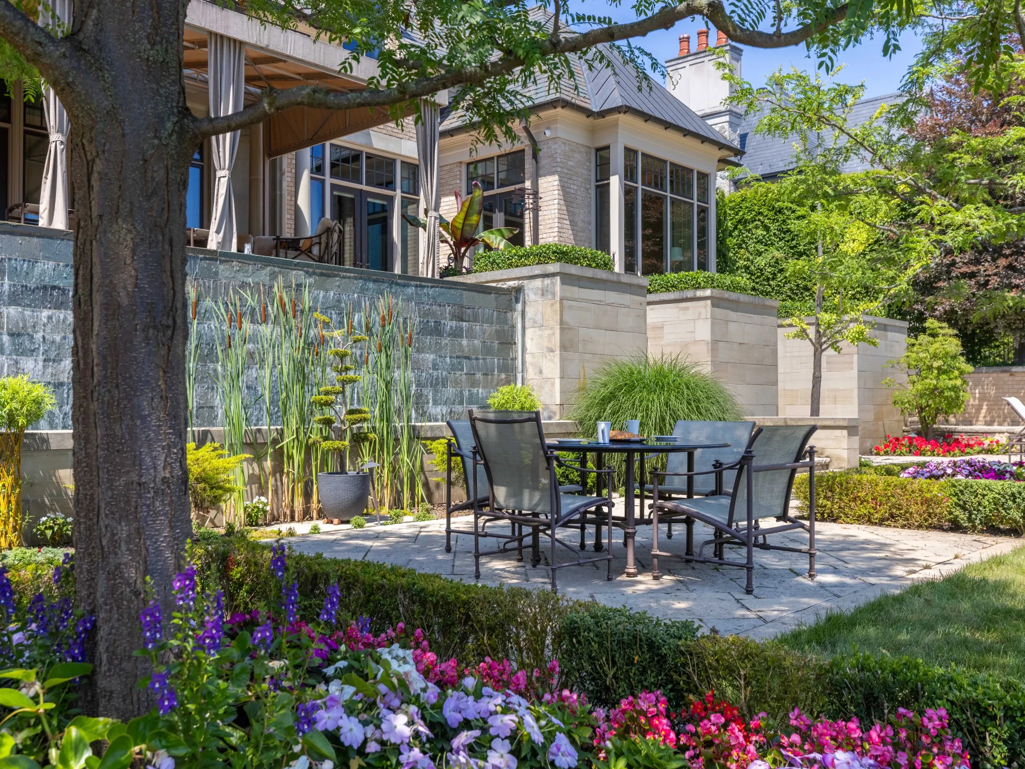 exterior patio with dining table, shaded by large trees and pretty pink flowers are in the garden