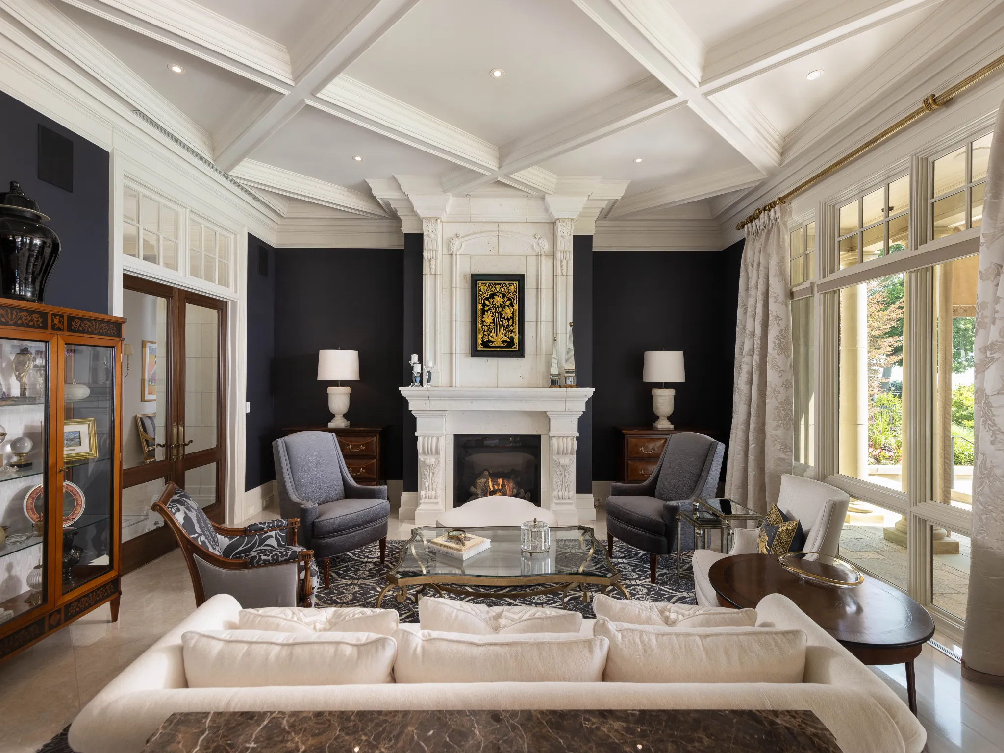 living room with navy walls and big windows. Large white fireplace and custom trim on ceiling.