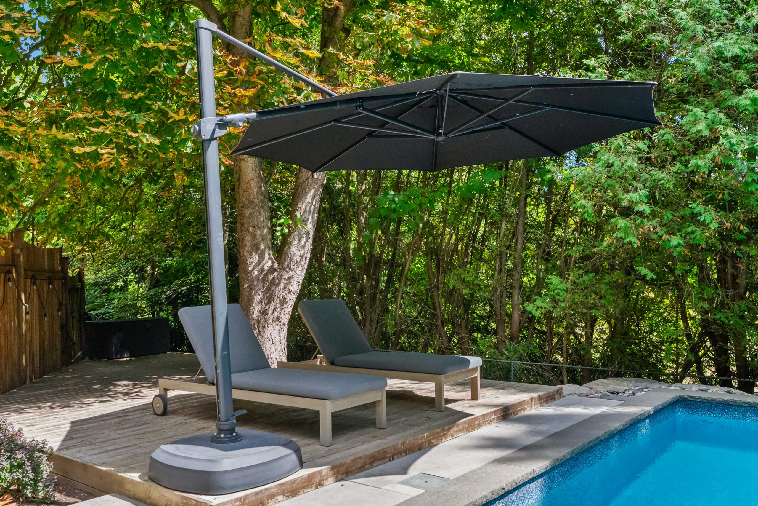 Black umbrella over two gray lounge chairs by a pool surrounded by greenery