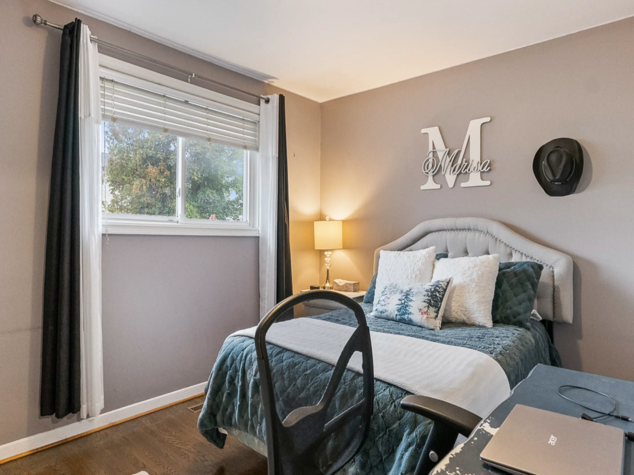 another bedroom with window and curtains with an m on the wall and lamp that is turned on