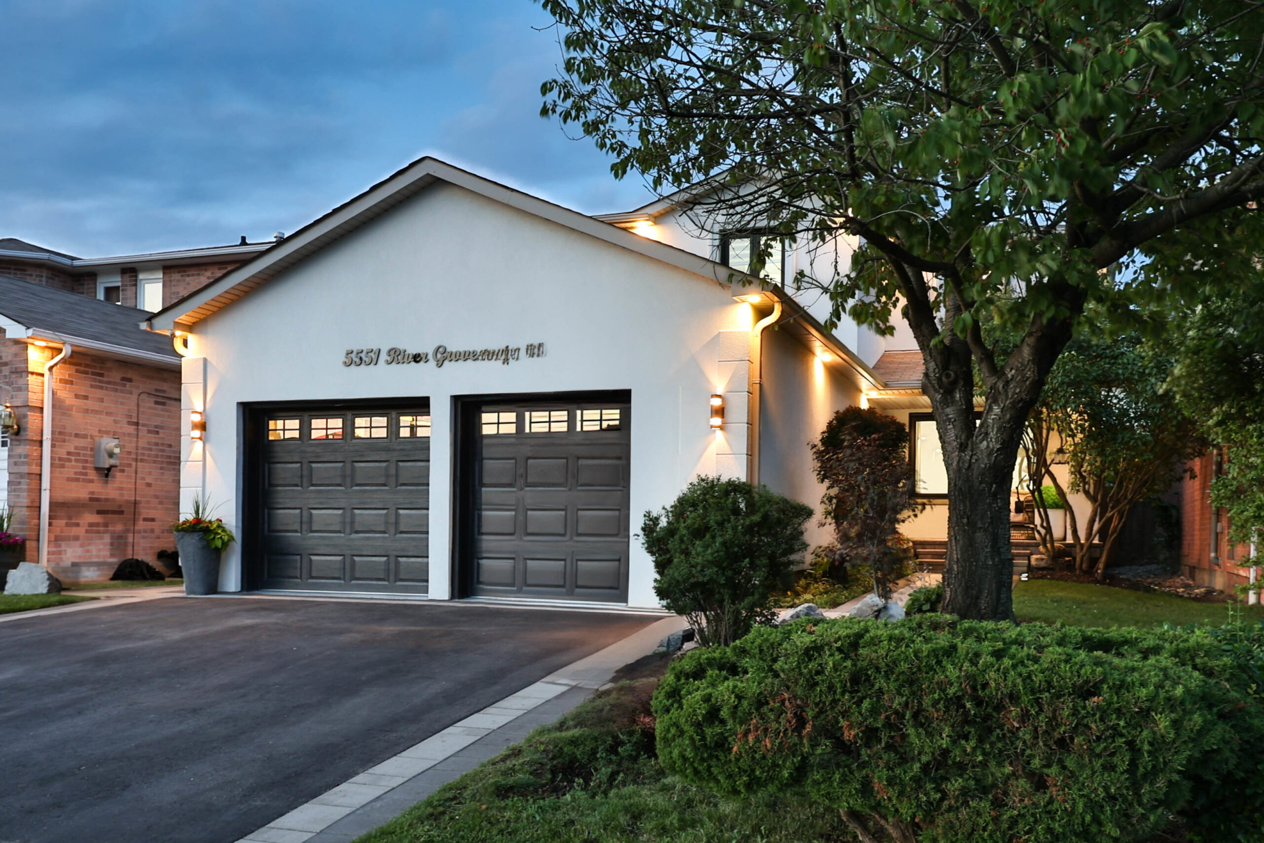 Modern house exterior with garage, illuminated at dusk, surrounded by greenery