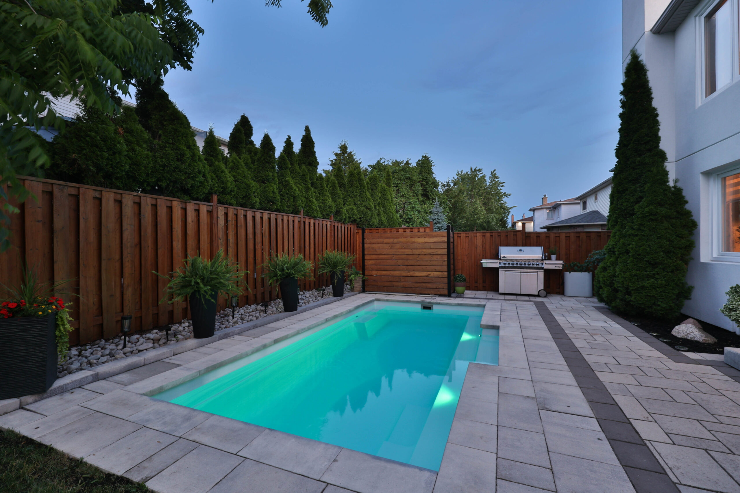Backyard pool area with illuminated water, stone patio, and wooden fence