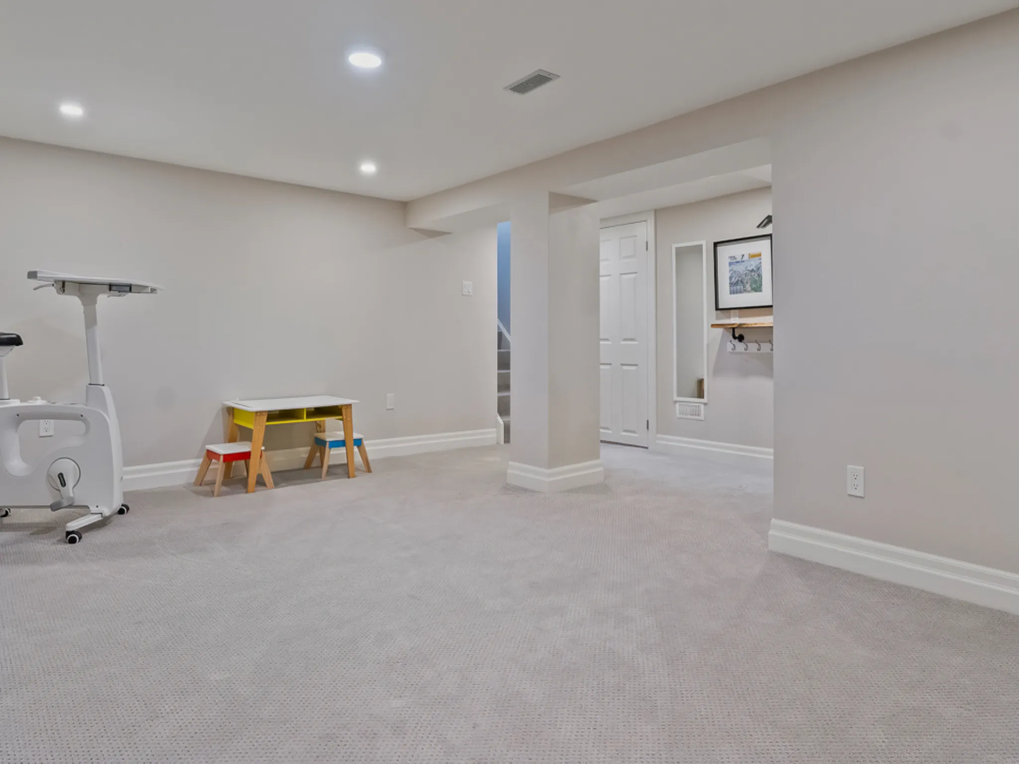 Bright basement room with carpet, exercise equipment, and colorful table.