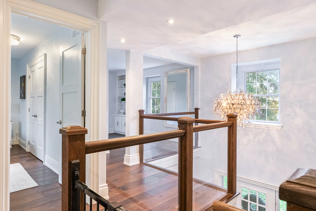 Bright hallway with wooden railing and modern chandelier, featuring light walls and hardwood floor