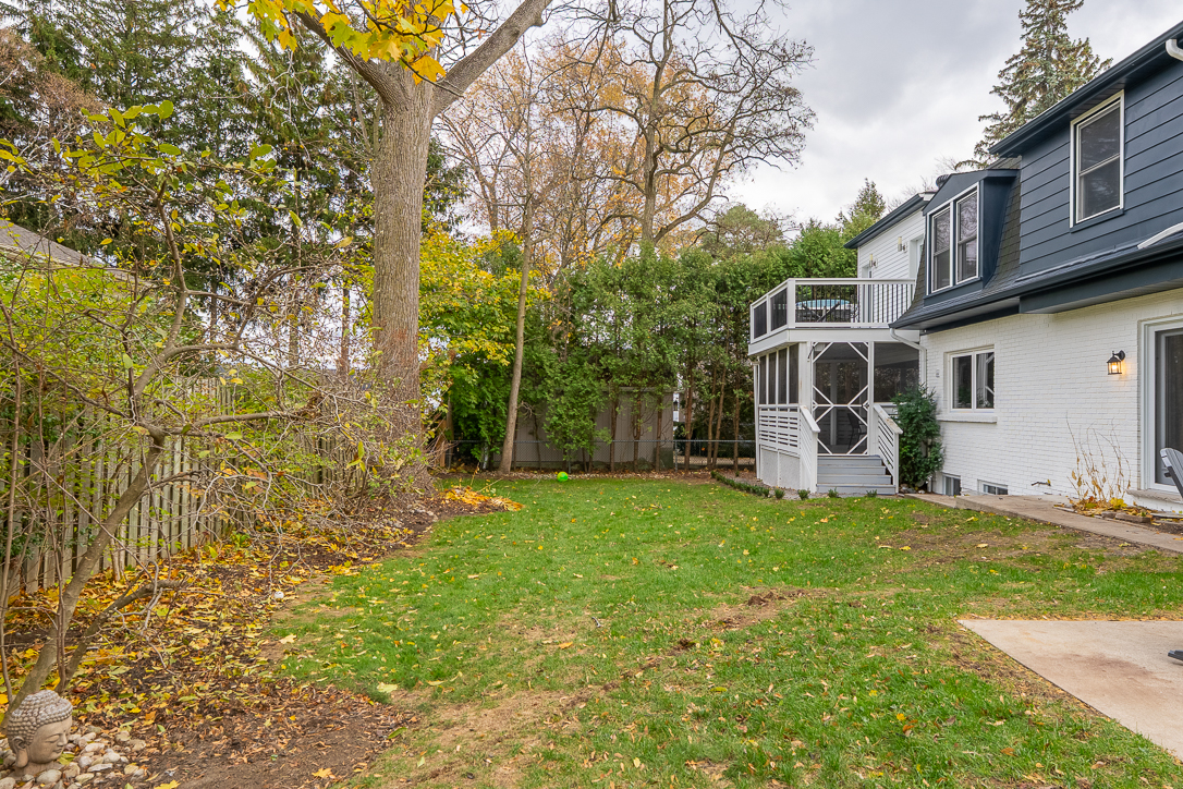 Backyard view featuring green lawn, trees, and a white house with a deck