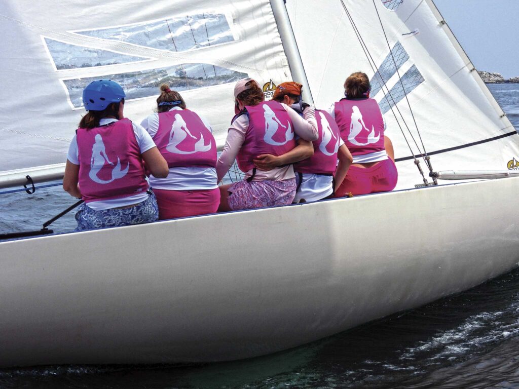 Five people sitting on a sailing boat all wearing pink lifejackets with a mermaid pictued on the jackets.