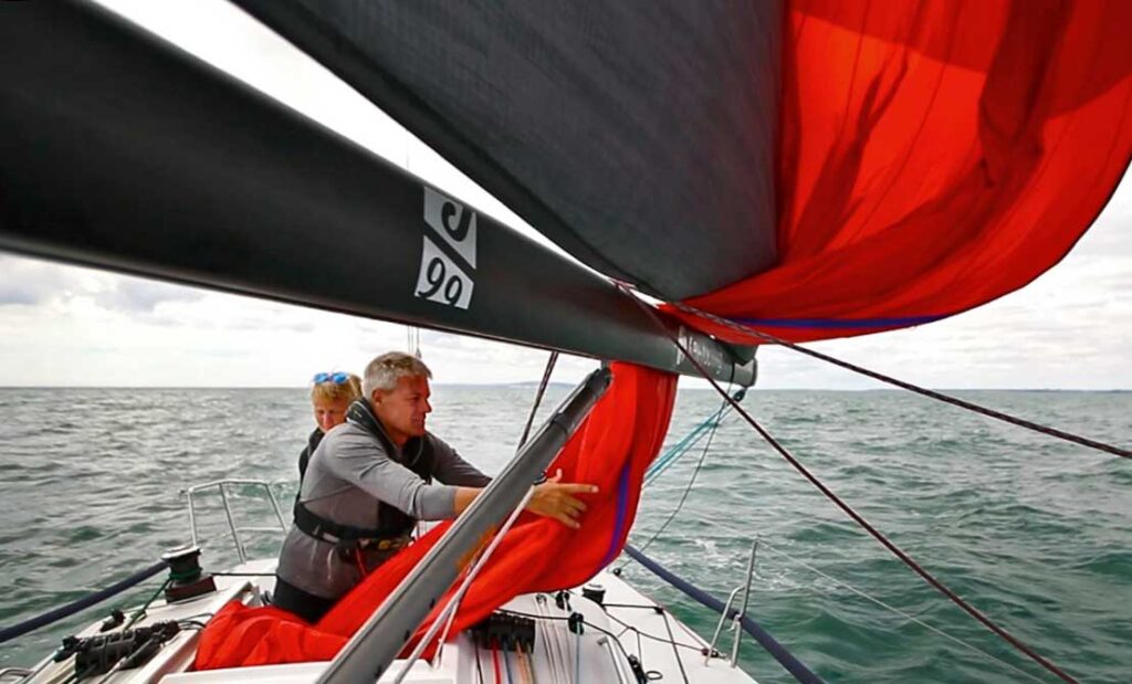A man and a woman on a boat in the water. The man is handling a red spinmaker.