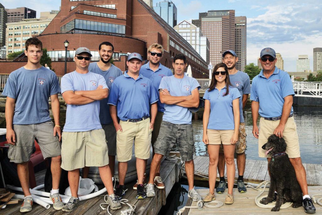 A group of people and a cute doggie standing on a dock. The dog is sitting like a good boy.