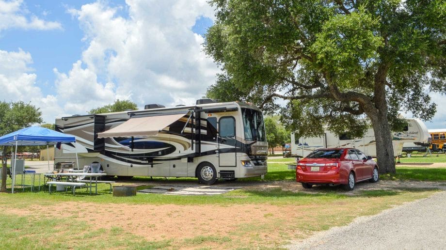 A large motorhome is parked at a campsite near a tree, with a red car parked beside it.