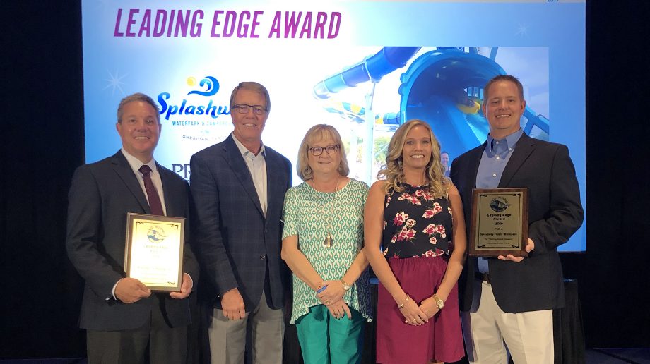 A group of five people is standing together. Two people are holding plaques, and a sign above them reads "Leading Edge Award." A water park image is in the background.