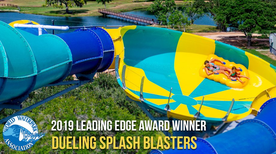 Two people riding a yellow inflatable raft on a bright yellow and teal water slide at a water park. The slide is surrounded by greenery and water. “2019 Leading Edge Award Winner Dueling Splash Blasters” text is displayed.