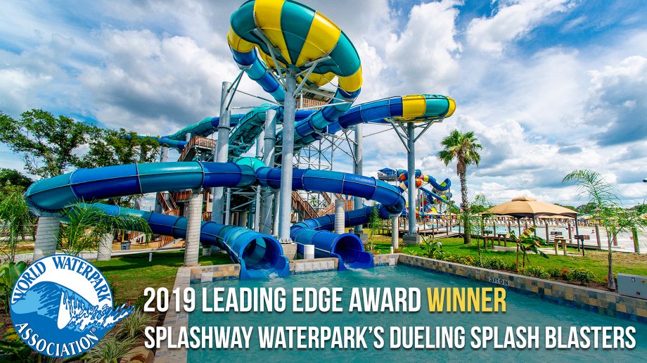 Two people riding a yellow inflatable raft on a bright yellow and teal water slide at a water park. The slide is surrounded by greenery and water. “2019 Leading Edge Award Winner Dueling Splash Blasters” text is displayed.