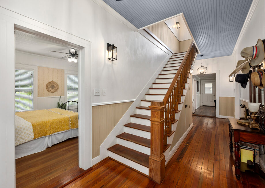 A hallway with wooden floors and a staircase. There is a doorway on the left leading to a bedroom with a yellow bedspread. Hats are hanging on the wall to the right. The ceiling is painted blue.