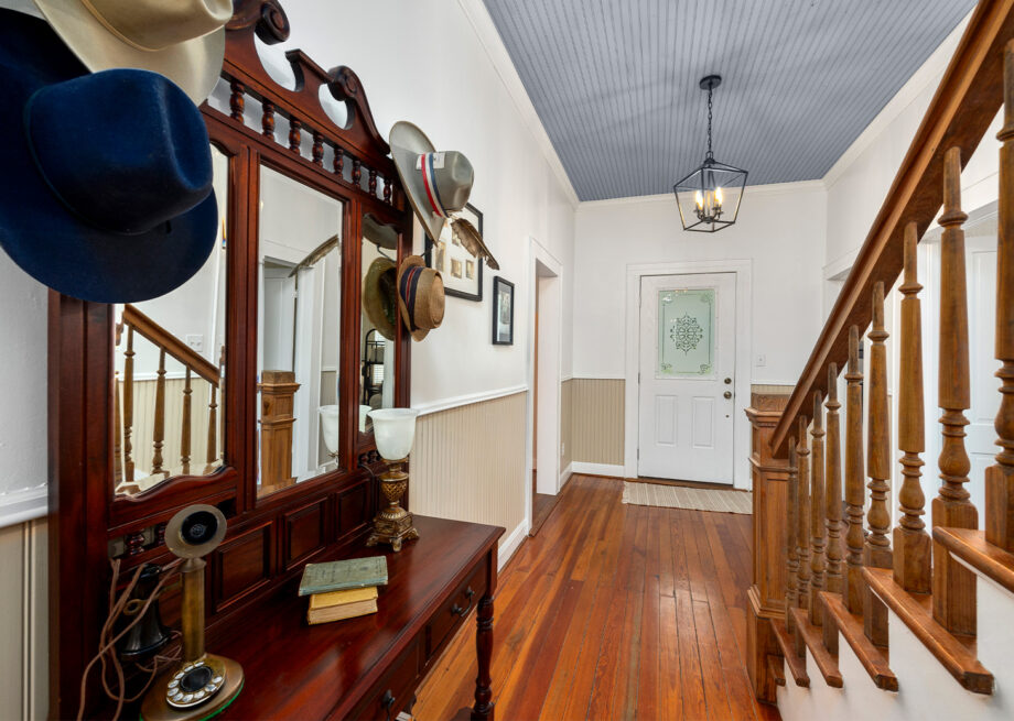 A hallway with wooden floors featuring a staircase, a mirror with various hats hanging on it, a vintage phone, and books on a wooden table.