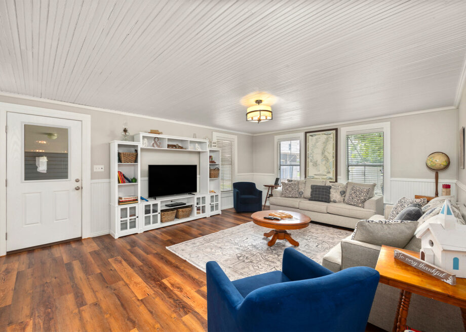 A cozy living room with hardwood floors, a white paneled ceiling, and a large area rug. It has a couch, two blue armchairs, a coffee table, and a white entertainment center with a mounted TV.