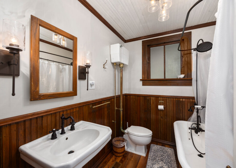 A bathroom with wood paneling, a wall-mounted sink, a toilet, a clawfoot bathtub with a curtain, and vintage-style fixtures.