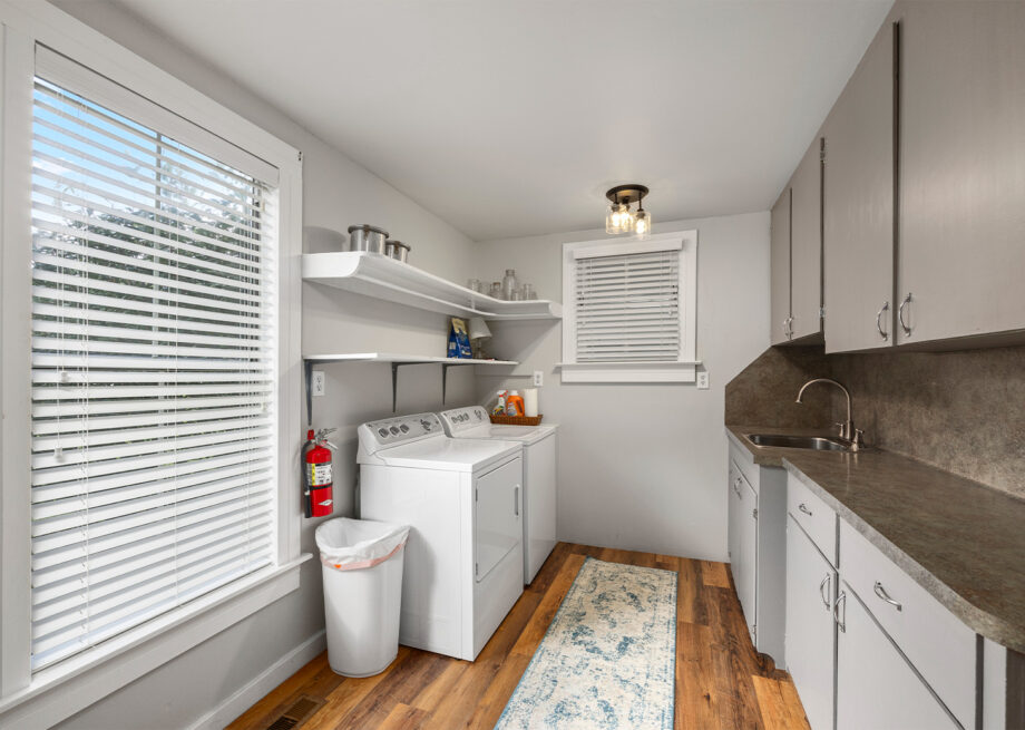 A laundry room with a washing machine, dryer, shelves, and cabinets. A fire extinguisher and trash can are next to the appliances. A rug lies on the wooden floor.