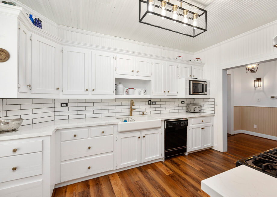 A kitchen with white cabinets, a white subway tile backsplash, a farmhouse sink, wood floors, and modern lighting.