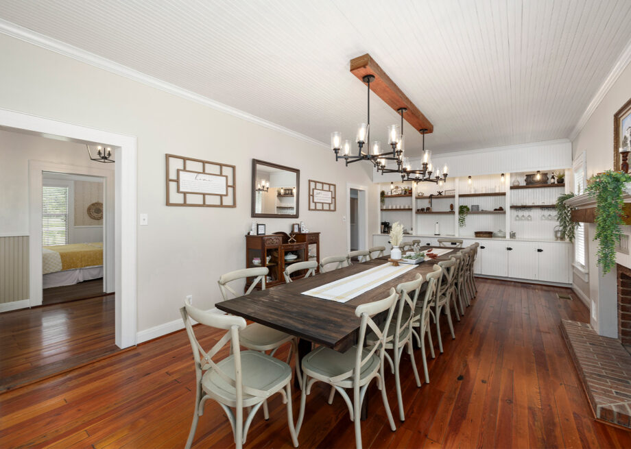 A spacious dining room with a long wooden table, numerous chairs, a chandelier, hardwood floors, and an open doorway leading to a bedroom.
