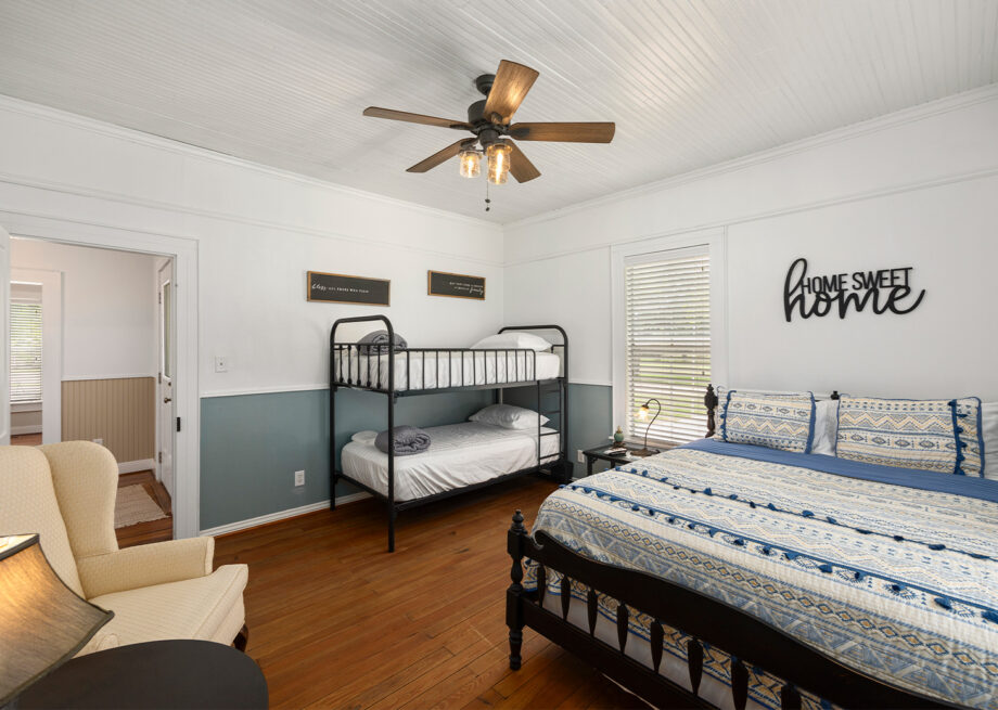 A bedroom with a queen bed, a bunk bed, a white armchair, a ceiling fan, and wall decor including "Home Sweet Home" sign.
