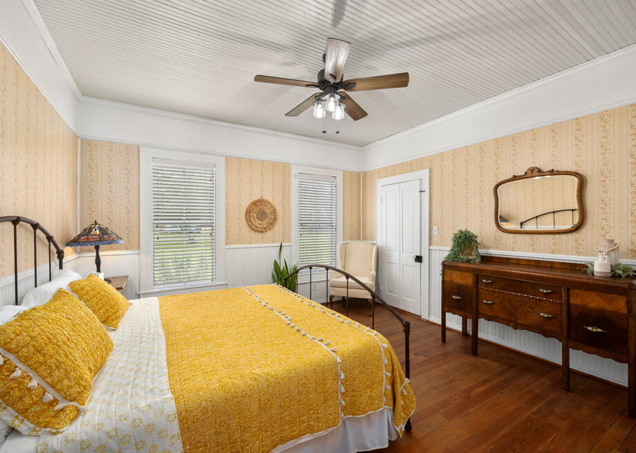 A bedroom with a yellow bedspread, wooden floor, and vintage furniture, including a dresser with a mirror above it. The room has white walls with yellow patterned wallpaper and a ceiling fan.