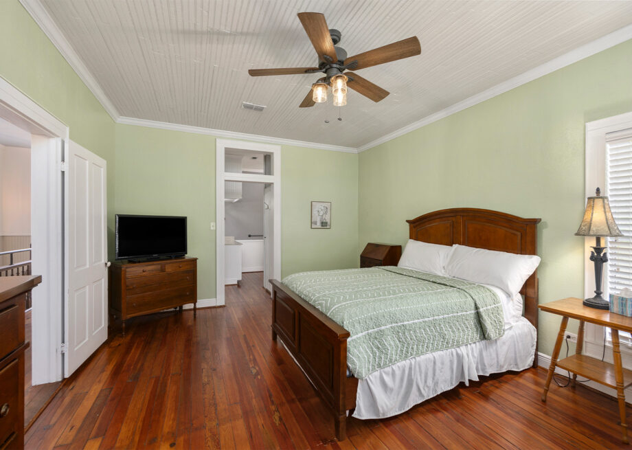 A bedroom with a wooden floor and green walls featuring a bed with green bedspread, ceiling fan, wooden furniture, a TV on a dresser, a lamp on a nightstand, and a doorway leading to an adjacent room.