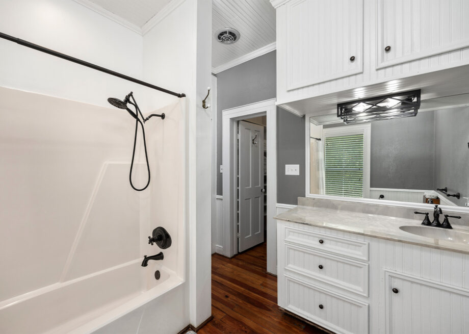 A bathroom with a white bathtub and black fixtures, a large mirror above a white countertop, white cabinetry, and wooden flooring.