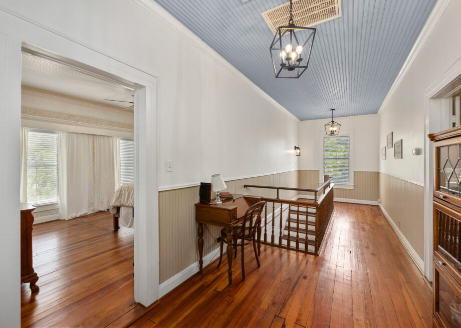 A hallway with wooden flooring, a small desk with a chair, and overhead light fixtures. The hallway leads to a bedroom with large windows and white curtains.