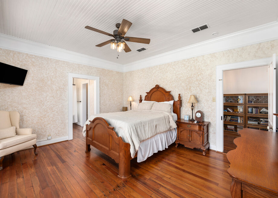 A bedroom with a wooden bed, white linens, a ceiling fan with lights, a wall-mounted TV, an upholstered chair, wooden nightstands, and an open door leading to a room with bookshelves.