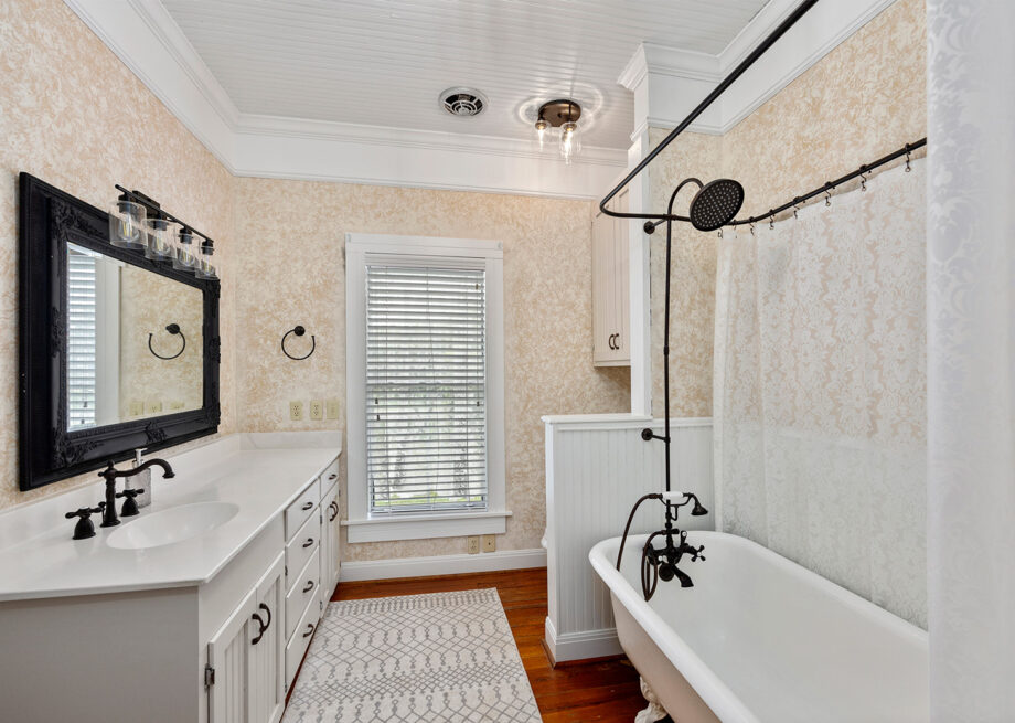 A bathroom with a clawfoot bathtub, shower curtain, double vanity with black-framed mirror, and a window with blinds.