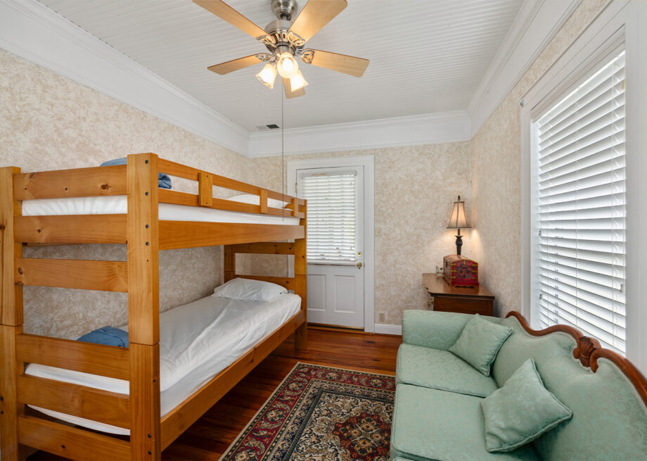 A bedroom with a wooden bunk bed, a light green couch, a rug, a door, and a window with blinds. A ceiling fan with lights hangs from the white ceiling. A lamp is on a side table next to the couch.