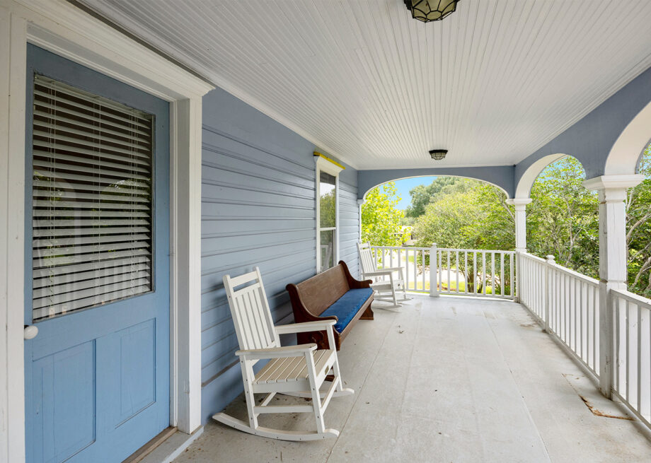 A spacious porch with light blue walls, a blue door, white rocking chairs, a wooden bench, and white railings.