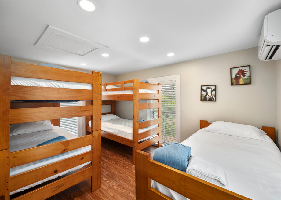 A room with two wooden bunk beds and one single bed, all made with white linens. There are framed pictures on the wall and an air conditioning unit mounted near the ceiling.