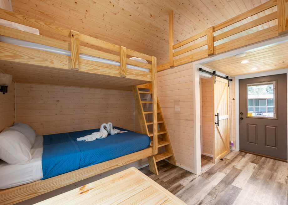 A small wooden cabin interior with bunk beds, a ladder, a sliding door, and a grey exterior door. The cabin has a rustic, minimalist design.