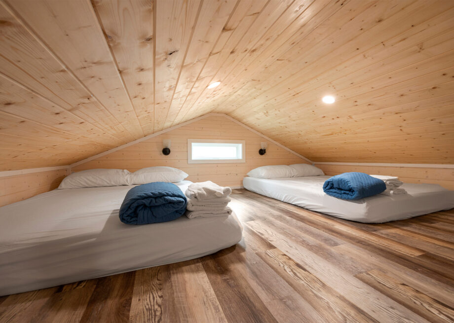 A small attic room with a wooden ceiling and floor, featuring two low beds with white sheets and blue blankets, each accompanied by folded towels.