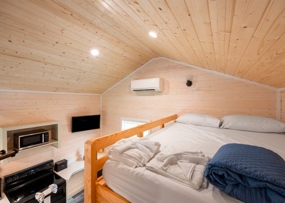 A cozy loft bedroom with wooden walls and ceiling, featuring a bed, microwave, small TV, stove, and air conditioning unit.