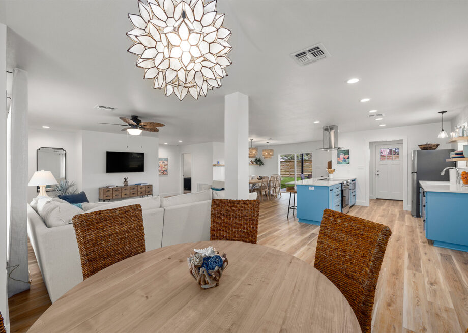 Open-concept living room and kitchen with light wood flooring, wicker dining chairs, white walls, a large round dining table, and blue accents in the kitchen.