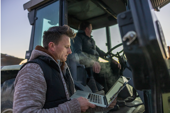 Man using a laptop in a tractor, another person driving in the background