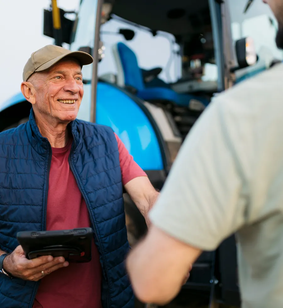 Smiling older man in a vest holding a tablet near a blue tractor