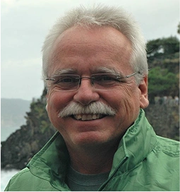 Smiling man with glasses wearing a green jacket outdoors near the coast