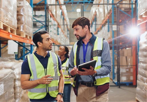 Two men in safety vests discussing in a warehouse aisle with shelves of goods
