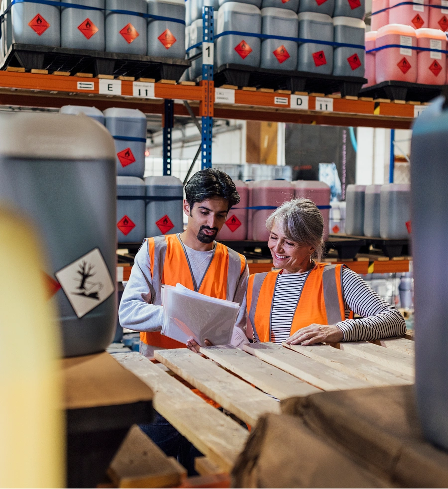 Two warehouse workers in safety vests reviewing documents among storage containers