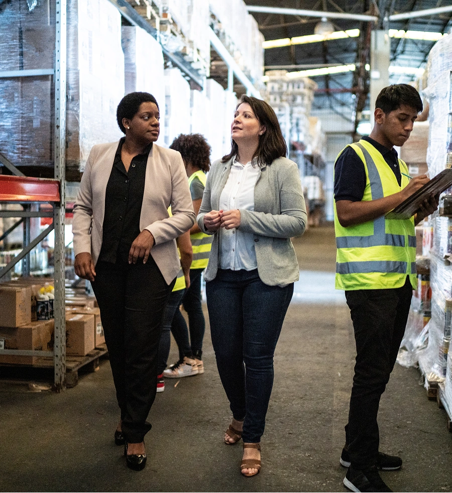 Two women walk through a warehouse while a man checks inventory on a tablet