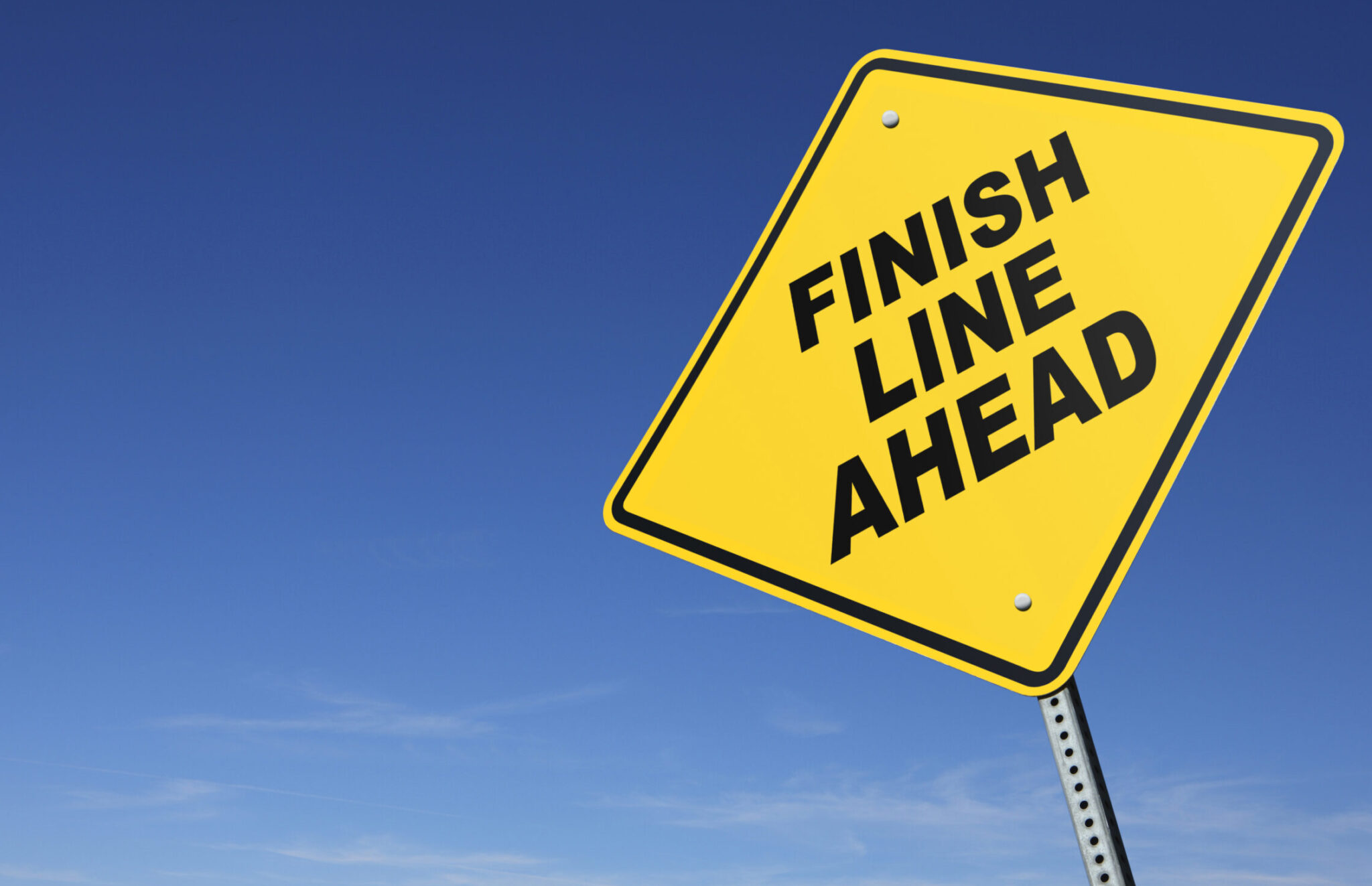 A yellow road sign warning that the finish line is ahead. A clear blue sky with wispy clouds at the horizon dominate the scene. Perspective is from a low camera angle. Road sign is placed on right side of image allowing for copy space on the left side of image.