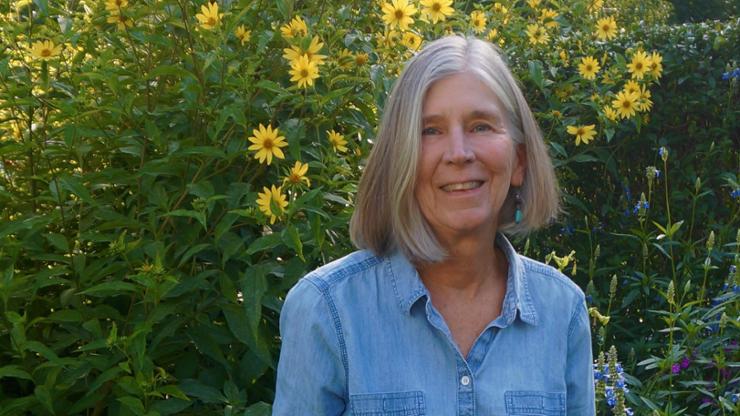 head and shoulders portrait of woman with grey hair and blue long-sleeve shirt standing in front of yellow flowers