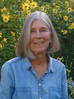 head and shoulders portrait of woman with grey hair and blue long-sleeve shirt standing in front of yellow flowers