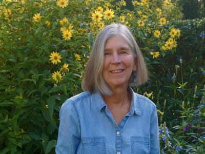 head and shoulders portrait of woman with grey hair and blue long-sleeve shirt standing in front of yellow flowers
